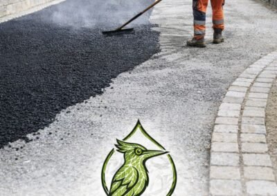 Ouvrier réalisant la pose d’un enrobé à chaud pour une cour extérieure avec bordures en pavés et fondation en grave ciment, logo Woodpecker Paysagiste en bas d’image
