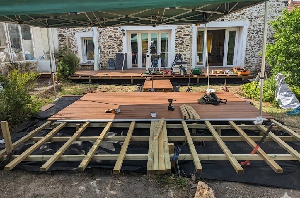 Image de chantier de construction de terrasse composite en Seine-et-Marne. Structure en lambourdes de bois visibles, deck fini sous tonnelle devant maison en pierre, outils de pose (visseuse, vis Spax).